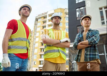 Drei seriöse, gutaussehende Arbeiter in Hardhats, die in die Ferne starren Stockfoto