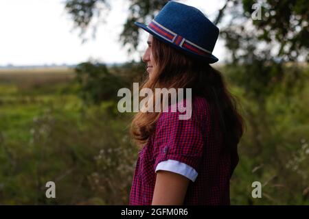 Unschärfe-Nahaufnahme Porträt einer lächelnden jungen Frau mit braunen Haaren, die einen Hut im Freien trägt. Grüne Natur Hintergrund. Rückansicht. Nicht fokussiert. Stockfoto