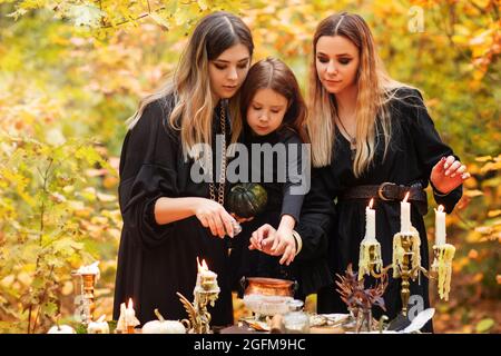 Glückliche Frauen und kleine Mädchen zerkleinern Kräuter in Mörtel während der Vorbereitung Zaubertrank während der Halloween-Feier am Herbsttag im Wald Stockfoto