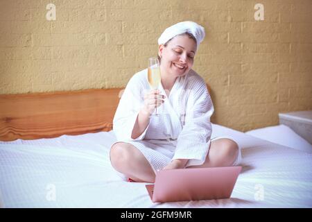 Die junge Hipster-Frau lächelt, während sie während eines Videoanrufs über einen Laptop zu Hause auf dem Bett sitzt, Champagner trinkt Stockfoto
