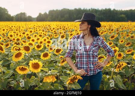 Die junge Agronomin trägt Cowboy-Hut, kariertes Hemd und Jeans auf dem Sonnenblumenfeld. Sommerliches Erntekonzept Stockfoto