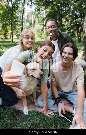 Positive multiethnische Teenager mit Retriever, die Selfie auf dem Handy im Park machen Stockfoto