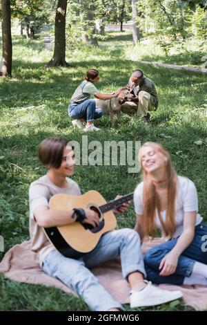 Interracial Teenager streicheln Retriever in der Nähe verschwommener Freunde spielen akustische Gitarre auf Rasen Stockfoto