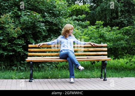 Allein eine junge Frau, die in Hemd und Jeans gekleidet ist und auf einer Bank im Park sitzt Stockfoto