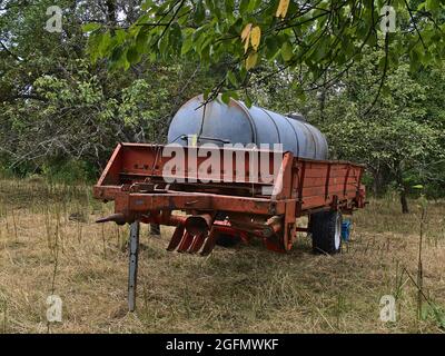 Blick auf einen alten, rostigen Tankwagen mit roten und grauen Farben, der auf einer Lichtung im Wald mit braunem Gras und grünen Bäumen in der Nähe des Limburger Hügels steht. Stockfoto