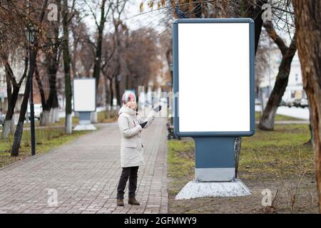 Die junge Frau zeigt auf eine leere vertikale Plakatwand auf der Gasse im Stadtpark. Leeres, leeres weißes Modell der Plakatwand. Copyspace. Stockfoto