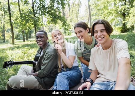 Happy teen girl sitzt auf Gras in der Nähe von multiethnischen Freunden mit Akustik-Gitarre Stockfoto