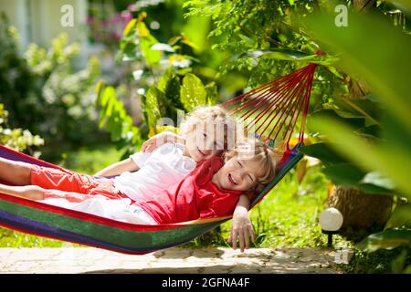 Kinder entspannen sich in einer bunten Regenbogenhängematte. Heißer Tag Garten Spaß im Freien. Junge spielt draußen. Brüder spielen draußen im sonnigen Hinterhof. Mittagsschlaf Stockfoto