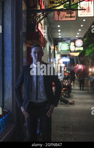 Das Porträt eines jungen, eleganten und reichen Geschäftsmannes in elegantem Anzug steht auf der Straße mit Lichtern in der Großstadt. Nachtbild. Neonschilder Stockfoto