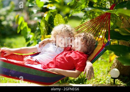 Kinder entspannen sich in einer bunten Regenbogenhängematte. Heißer Tag Garten Spaß im Freien. Junge spielt draußen. Brüder spielen draußen im sonnigen Hinterhof. Mittagsschlaf Stockfoto