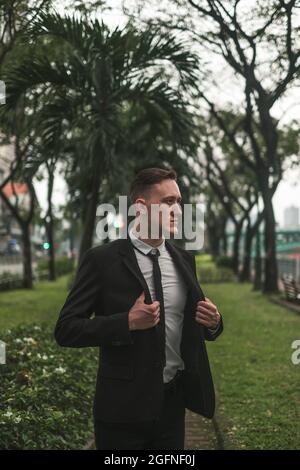 Porträt eines hübschen kaukasischen jungen Geschäftsmanns in schwarzem eleganten Anzug, der im Park spazieren geht. Blick beiseite. Seriöse Optik. Sommerzeit. Stockfoto