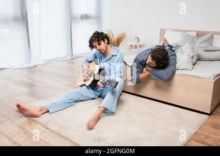 gay Mann spielt akustische Gitarre in der Nähe Freund auf dem Bett Stockfoto