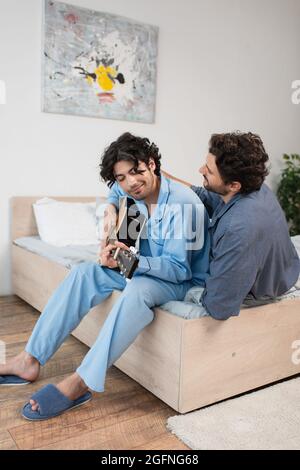 Glücklich schwulen Mann spielt akustische Gitarre in der Nähe Freund auf dem Bett Stockfoto