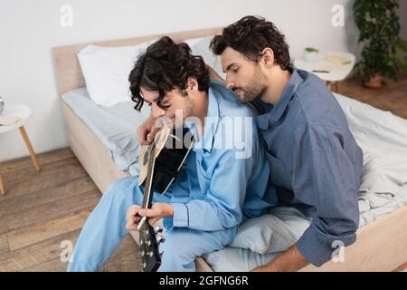 gay Mann spielt akustische Gitarre in der Nähe bärtigen Freund auf dem Bett Stockfoto