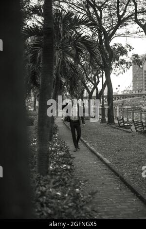 Hübscher junger Kaukasier in schwarzem Anzug und Sonnenbrille, der im Park spazieren geht. Park mit tropischen Pflanzen. Schwarzweiß-Foto Stockfoto