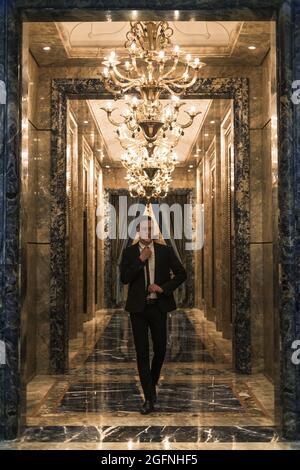 Ein junger, hübscher kaukasischer Geschäftsmann in elegantem Geschäftsanzug, der mit wunderschönen, teuren Kronleuchtern auf dem Flur herumläuft. Richtet seine Krawatte aus Stockfoto