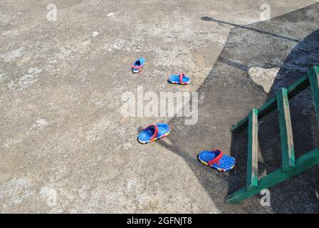 In der Nähe einer Holztreppe auf einem Trampolin auf einem Betonboden sind zwei Paar farbige Kindersandalen verstreut Stockfoto