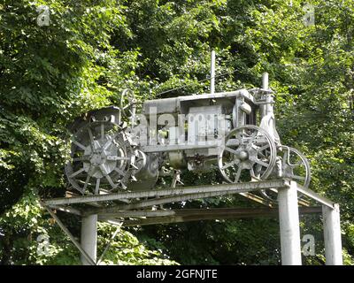 Ein sehr alter Traktor, der außer Betrieb ist, steht auf einem Sockel, wie ein Denkmal auf dem Hintergrund von Bäumen im Park Stockfoto