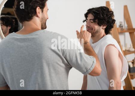 Tätowiert und verschwommen Mann Anwendung Creme auf die Nase des lächelnden Freundes Stockfoto