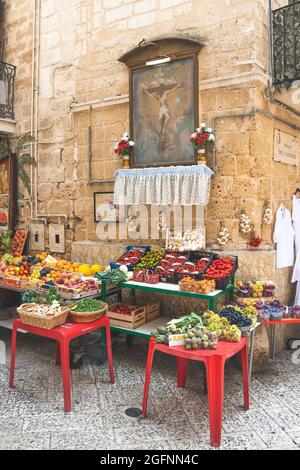 Blick auf eine schmale Straße in der Altstadt von Bari, Apulien, Italien, traditioneller offener Obstmarkt mit religiösen Bildern und verschiedenen lokalen Obst- und Gemüsesorten Stockfoto
