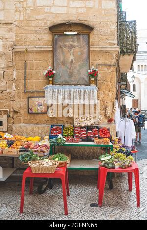 Blick auf eine schmale Straße in der Altstadt von Bari, Apulien, Italien, traditioneller offener Obstmarkt mit religiösen Bildern und verschiedenen lokalen Obst- und Gemüsesorten Stockfoto