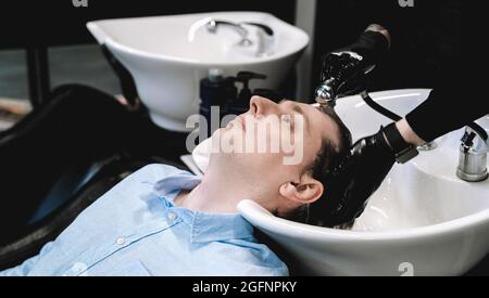 Besuch im Friseurladen. Junger stilvoller Mann, Geschäftsmann macht modische Frisur. Coiffeur, Friseur, Stylistin wäscht seine Haare mit Shampoo, Balsam in Stockfoto