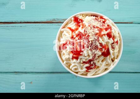 Serviert werden deutsche Vanillespaghetti-Eisbecher in einer Wanne zum Mitnehmen, die mit frischen Erdbeercoulis oder Sauce über blauem Holz mit Polizist gekrönt ist Stockfoto