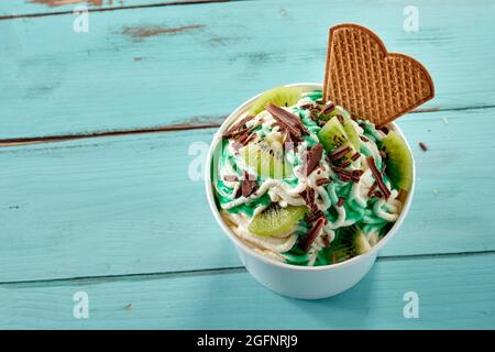 Tropische Kiwifruit auf einem trendigen deutschen Vanillespaghetti-Eisbecher, bestreut mit Schokoladenflocken und serviert mit einem Waffelbiskuit über blauem Woo Stockfoto