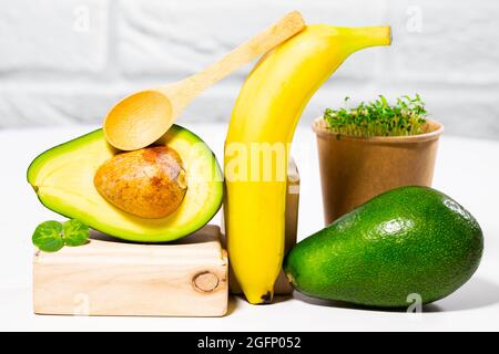 Kreatives Stillleben mit Avocado, Banane und Micro Green auf dem Hintergrund mit Holzständern und Podien. Gleichgewicht gesunde Lebensmittel. Ausgewogener Nutrit Stockfoto