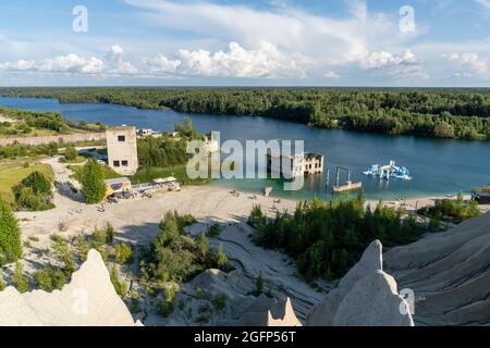 Rummu, Estland - 12. August 2021: Kalksteinberge und ruhiger blauer Grundwassersee in einem alten Steinbruch mit verlassenen Gebäuden im Wasser Stockfoto