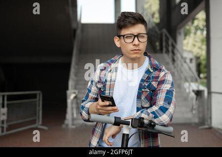 Kaukasischer Mann in Brille steht in der Nähe des Elektrorollers und hält sein Smartphone in den Händen. Stockfoto