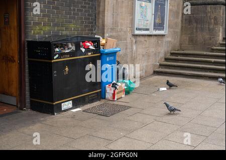 Im Stadtzentrum von Norwich gibt es Mülltonnen und blaue Mülltonnen, die mit Müll überfüllt sind und Tauben auf dem Bürgersteig neben einigen Stufen nach Futter suchen Stockfoto