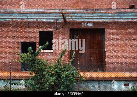Verrostete Fabriktüren in einer Ziegelwand auf einem verlassenen Laderampe mit einem Kettengliederzaun im Vordergrund Stockfoto