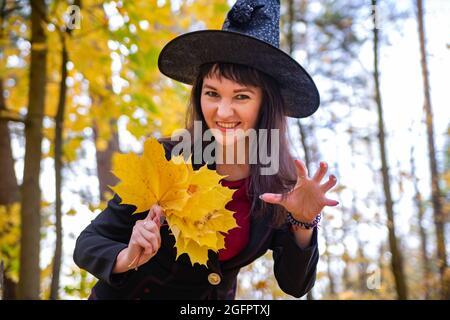 Lächelnde Frau in Form einer Hexe feiert halloween, in einem schwarzen großen Hut, Frau halten goldene Ahornblätter auf Herbst Wald Hintergrund Stockfoto