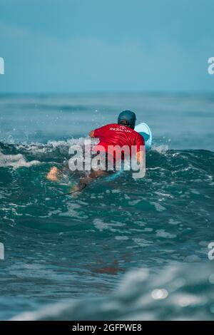 Playa Hermosa, Guanacaste, Costa Rica - 07.26.2020: Ein Mann mit rotem Hemd und blauer Mütze treibt langsam auf seinem Surfbrett zum Meer. Stockfoto