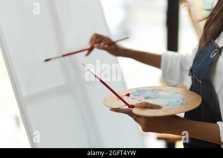 Beschnittenes Bild, junge Künstlerin hält Palette und malt im Studio mit dem Pinsel auf Leinwand Stockfoto