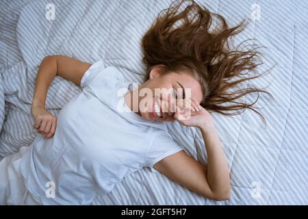 Schöne junge Frau, die lächelnd im Bett liegt. Ein Mädchen mit offenen Augen bedeckt morgens ihr Gesicht mit ihren Händen und Fingern. Happy Girl Konzept. Blick von oben. Die Freude des Erwachens Stockfoto