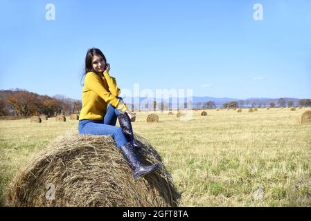 Junge Brünette Frau in gelben Pullover und Gummistiefel sitzen auf Heuhaufen im Feld Stockfoto