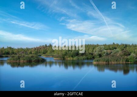 Ein See mit einer Ebenen Oberfläche und Reflexionen gegen den blauen Himmel und weiße Wolken. Entstanden aus einem alten Steinbruch überflutet, Gruppen von Inseln überwuchert Stockfoto