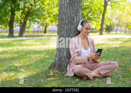 Foto einer glücklichen lächelnden 40-jährigen Frau, die Kopfhörer mit einem Smartphone trägt. Sie hört ihre Lieblingsmusik, während sie auf dem Gras im p sitzt Stockfoto