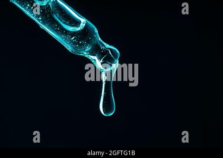 Pipette mit Flüssigkeit in Neonlicht. Auf dunklem Grund fällt ein Tropfen von einer Pipette. In Neonblau, Frontalansicht. Stockfoto