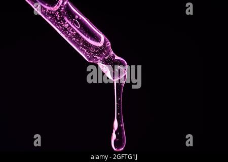 Pipette mit Flüssigkeit in Neonlicht. Auf dunklem Grund fällt ein Tropfen von einer Pipette. In Neonpink, Vorderansicht. Stockfoto