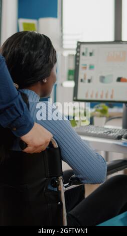 Ein Geschäftsmann hilft seinem behinderten schwarzen Kollegen, der in das Firmenbüro eintritt und den Rollstuhl in die Nähe des Schreibtisches schiebt. Afrikanisch behinderte gelähmte Frau begrüßt Kollegen, die bei einem Finanzprojekt arbeiten Stockfoto