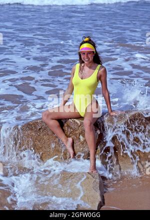 Archiv 1991 Ansicht eines lächelnden Badeanzug-Models, das auf Felsen in Meereswellen an einem heißen sonnigen Tag an der Küste an einem portugiesischen Sandstrand posiert, mit einer Spitzenmütze und einem gelben einteiligen Schwimmkostüm aus den 1990er Jahren, Archivbild, das in Portugal arbeitet Stockfoto