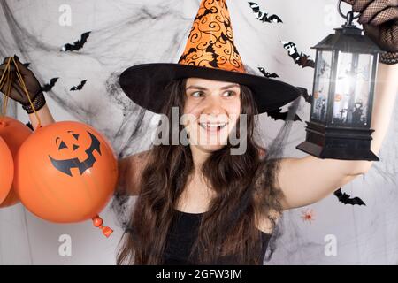 Eine Frau in einem Hexenkostüm mit Hut und Handschuhen im Hintergrund mit Fledermäusen hält Kürbiskugeln, glühende Laterne und lacht unheimlich. Halloween Stockfoto