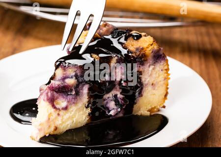 Mit einer Metallgabel, die einen Bissen von einem Stück hausgemachter Heidelbeere nimmt, und einem bröckeligen Käsekuchen, der mit Schokolade in weißer Keramikplatte auf einem Holztisch überzogen wird Stockfoto