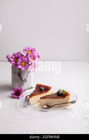 Scheiben selbstgebrannter baskischer Käsekuchen auf einem Teller mit Blaubeeren und Minzblättern auf hellem Hintergrund mit Blumen in Vase Stockfoto