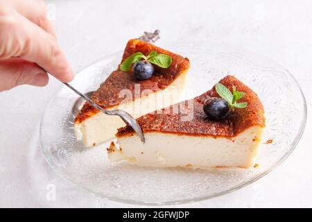 Die Hand einer Frau nimmt ein Stück Käsekuchen mit einem Löffel. Scheibe selbstgebrannter baskischer Käsekuchen mit Blaubeeren auf hellem Hintergrund. Stockfoto