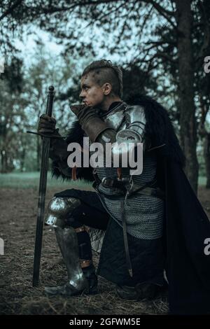 Mittelalterlicher Krieger-Ritter in Kettenpanzerung und schwarzem Umhang, der mit seinem Schwert auf einem Knie im Wald steht. Stockfoto