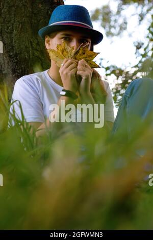 Unschärfe-Porträt einer lustigen jungen Frau mit braunen Haaren, die einen Hut im Freien trägt. Weibliche Hand mit gelben trockenen Blättern und bedecktem Gesicht. Hallo Herbst. Stockfoto